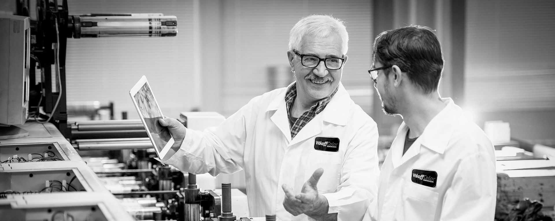 Two men in lab coats discuss while inspecting machinery; one holds a tablet, both appear engaged in conversation in an industrial setting.