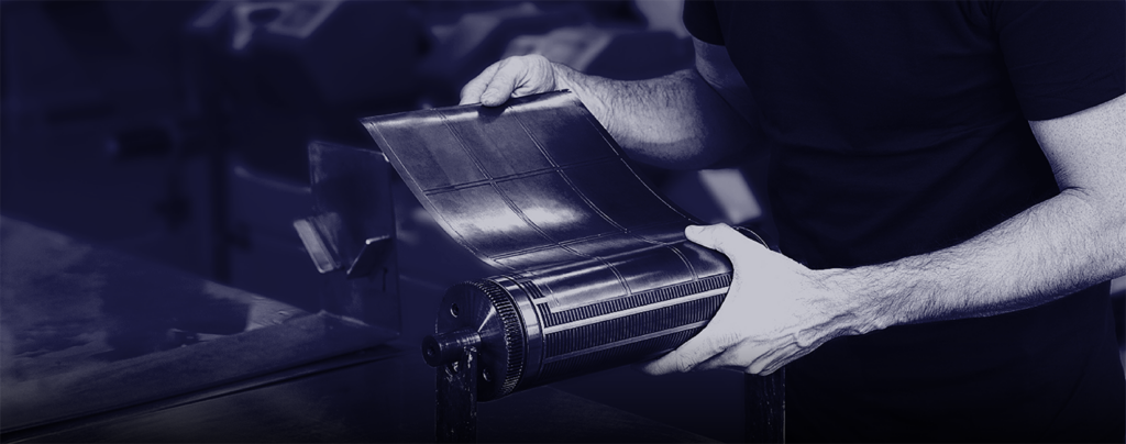 A person holds a flexible printing plate above a printing cylinder in an industrial setting.