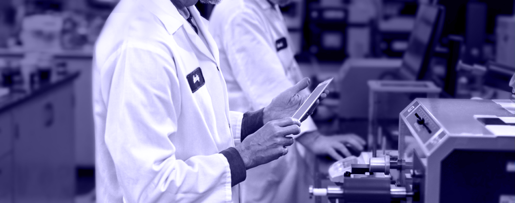 Two people in lab coats work with equipment and a tablet in a laboratory setting.