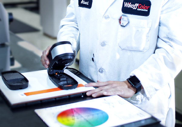 A person in a lab coat uses a handheld device to measure color from an orange strip, with a color chart and documents on the table.
