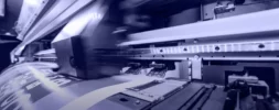 Close-up view of a large industrial printer in operation, producing printed material with visible mechanical components and movement.