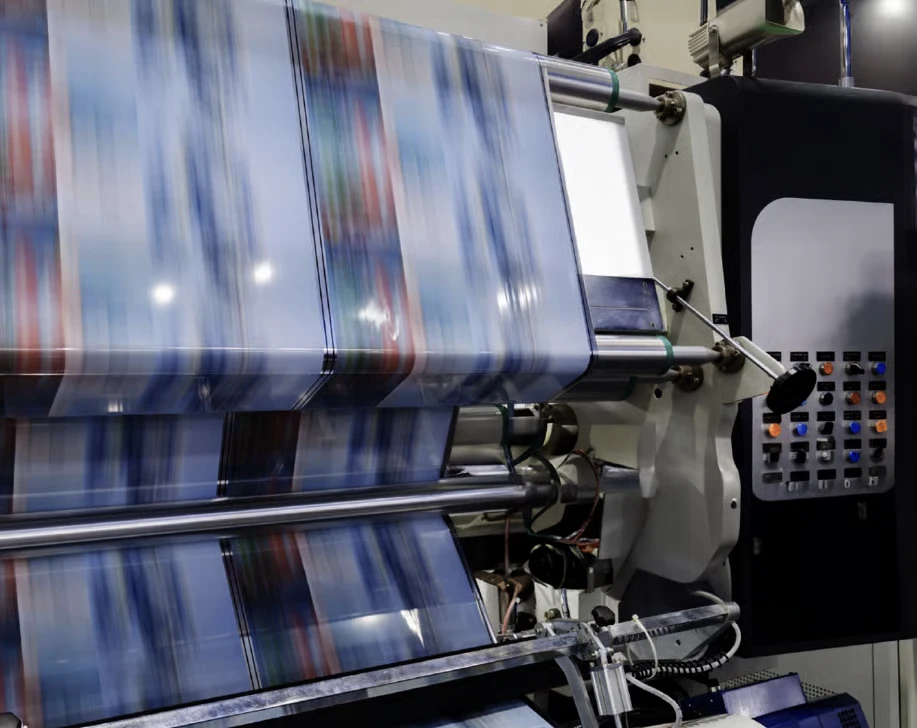 A close-up of a printing press in motion, displaying colorful rolls of printed material with machinery controls visible on the side.