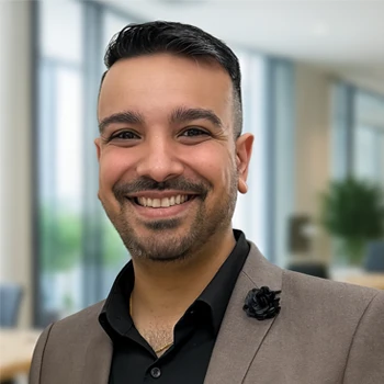 A man in a stylish brown blazer with a black flower pin stands confidently in a modern, bright office interior.