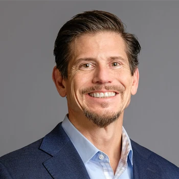 A man in a navy blazer with a light blue shirt poses against a gray background, exuding professionalism and confidence.
