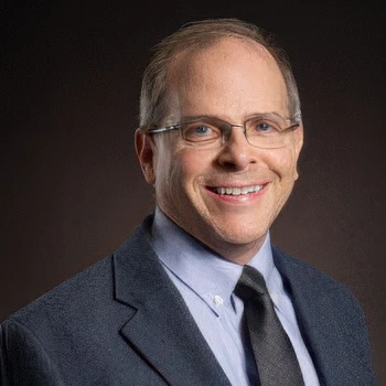 A professional man in a dark blazer and tie poses against a neutral backdrop, exuding confidence and approachability.
