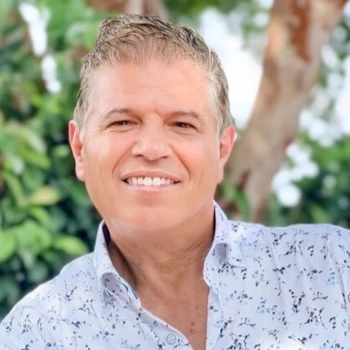 A man stands outdoors against a backdrop of lush green foliage, wearing a white shirt with a subtle floral pattern.