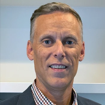 A man in a checked shirt and blazer stands in front of a softly blurred background, emphasizing a professional setting.