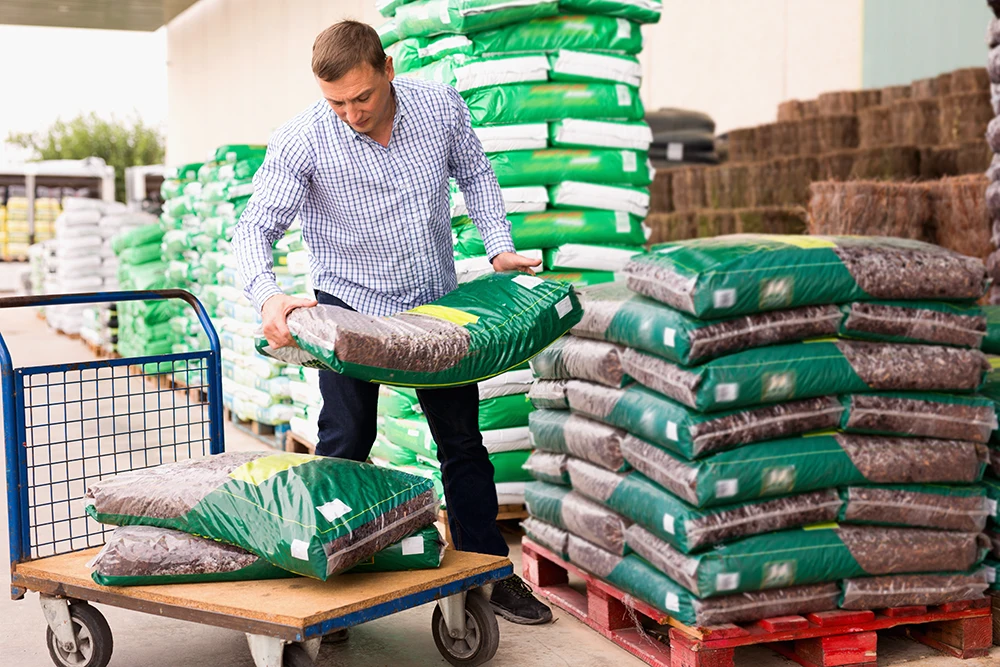 A man is lifting a large bag of soil onto a cart surrounded by stacks of similar bags in a garden supply store.
