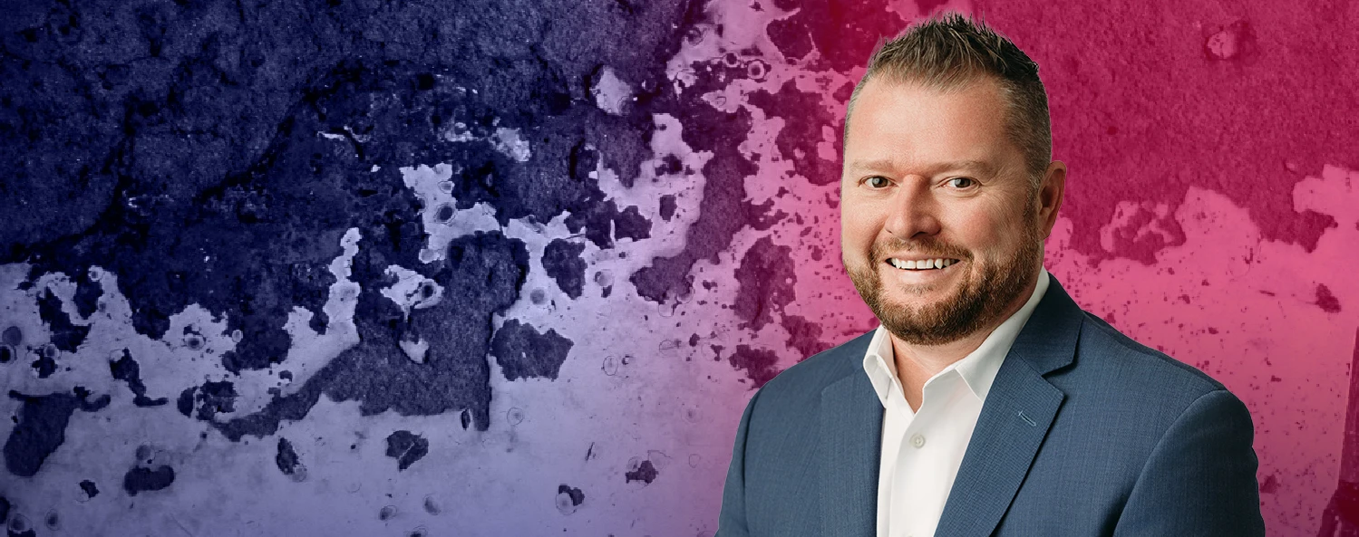 A man in a blue suit and white shirt smiles in front of a textured purple and pink background.