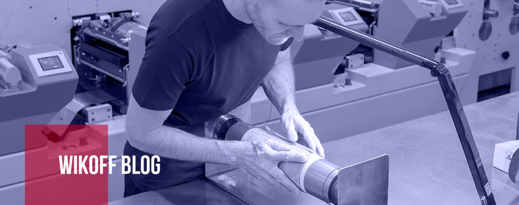 A man inspects a cylindrical printing component in an industrial setting with printing machines in the background; "Wikoff Blog" text is overlaid.