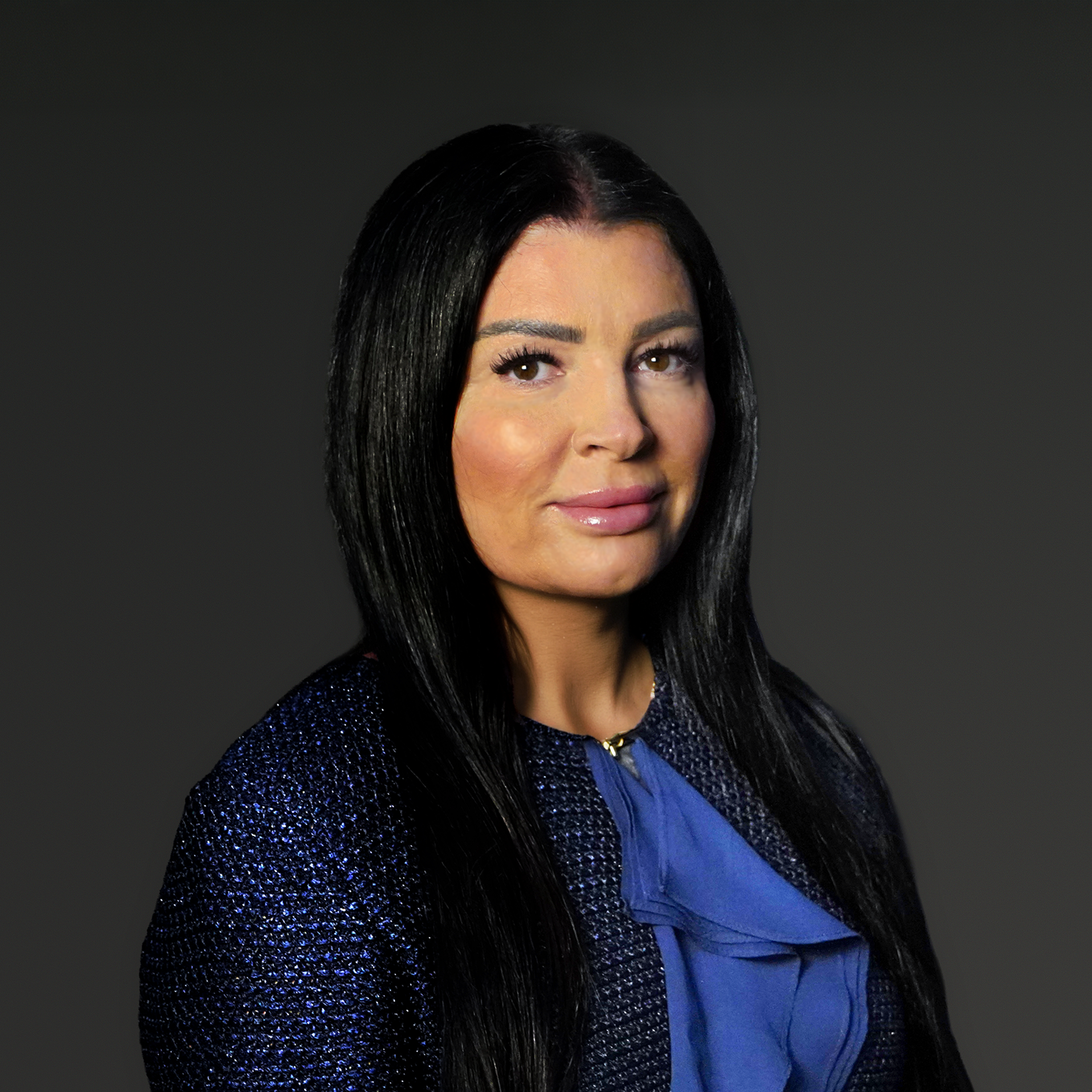 A woman with long straight black hair wearing a dark textured jacket and a blue blouse poses against a dark grey background.