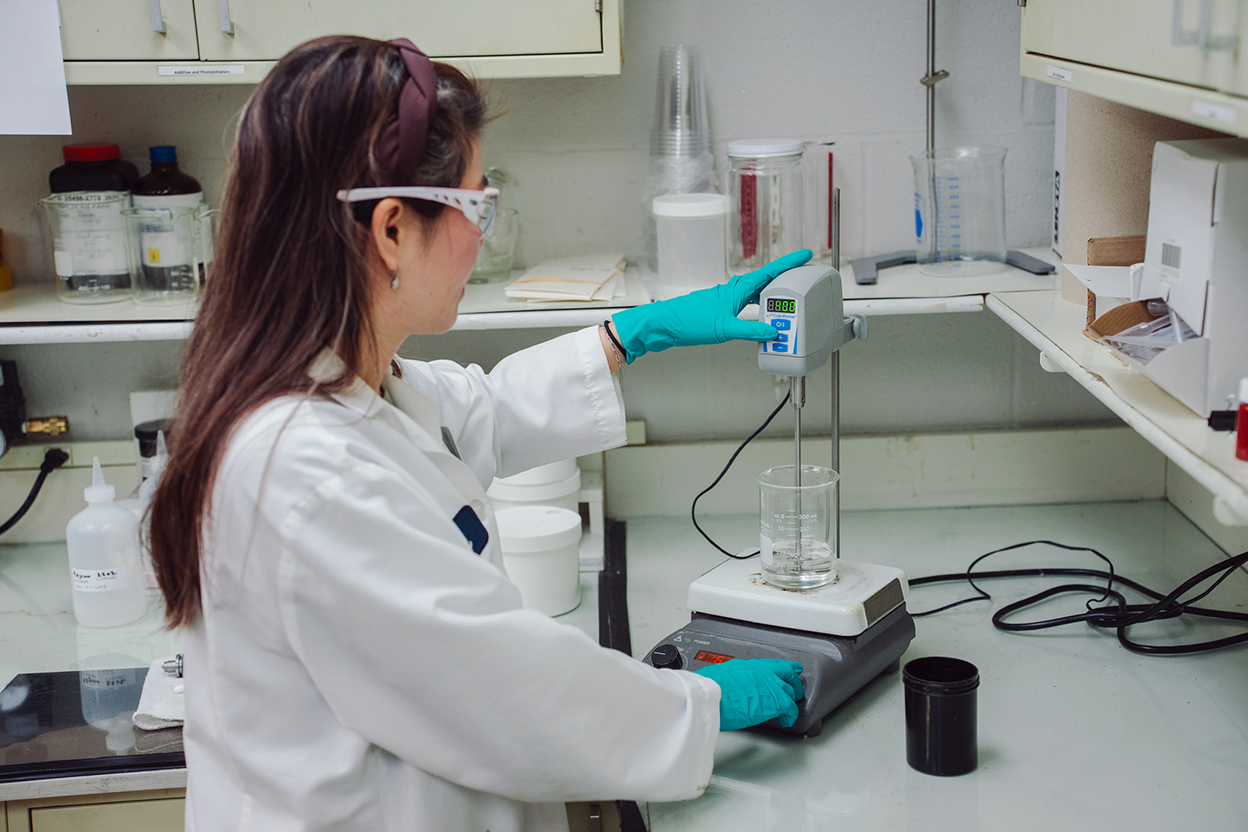 A person in a lab coat and gloves uses a digital mixer to stir a liquid in a glass beaker on a laboratory workbench.