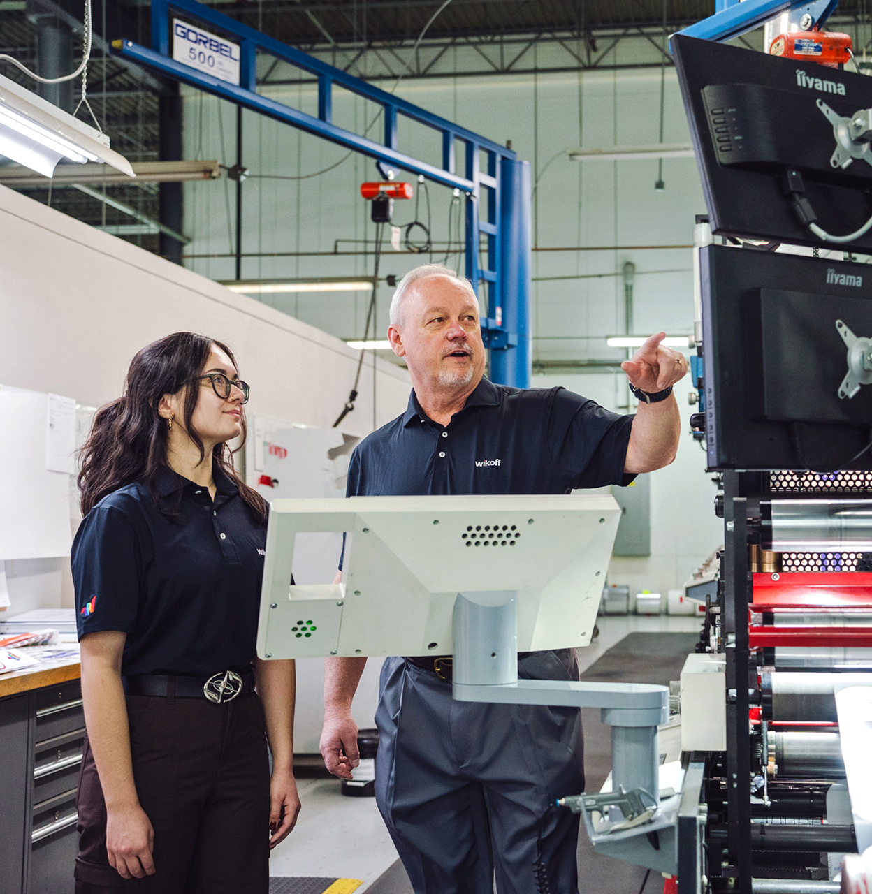 Two people in a factory setting stand next to computer monitors; one person points at the screens while the other looks on.