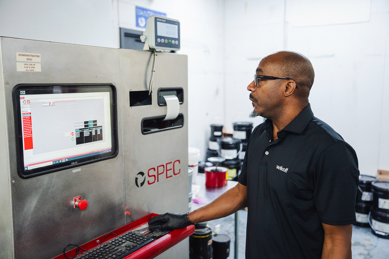 A man in a black polo shirt uses a computer at a SPEC industrial workstation in a lab with paint containers in the background.