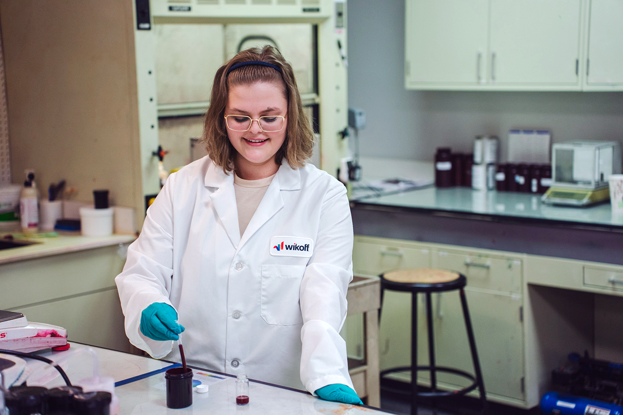 A person in a lab coat and gloves mixes a dark liquid in a small container at a laboratory workstation.