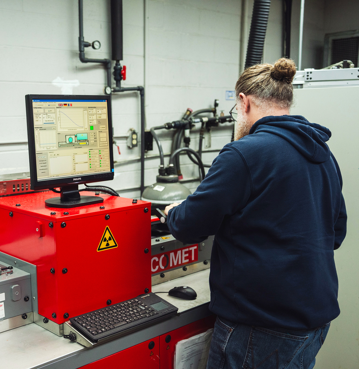 A person operates industrial equipment using a computer interface in a lab setting, with safety and monitoring devices visible around the workstation.