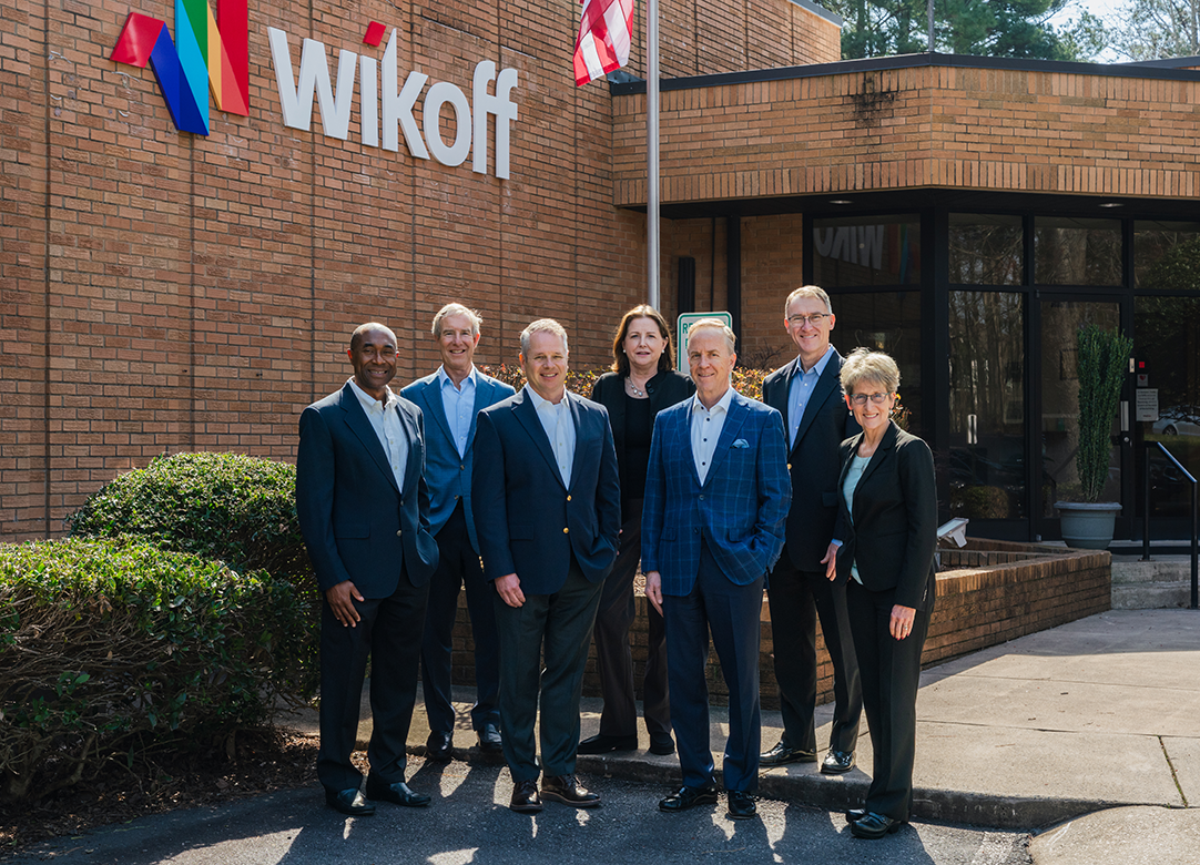 Seven people in business attire stand outside the Wikoff building next to an American flag, posing for a group photo on a sunny day.