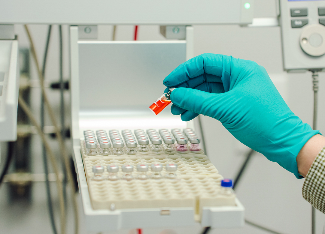 A gloved hand holds a small vial of red liquid over a tray of sample vials in a laboratory setting.