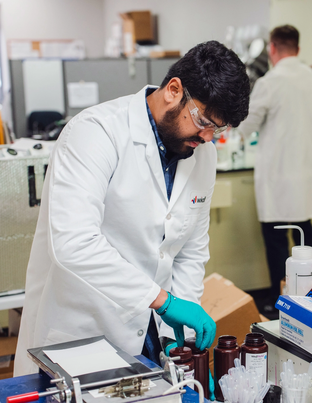 A man in a lab coat and gloves handles brown bottles at a laboratory workstation, with lab equipment and another person in the background.