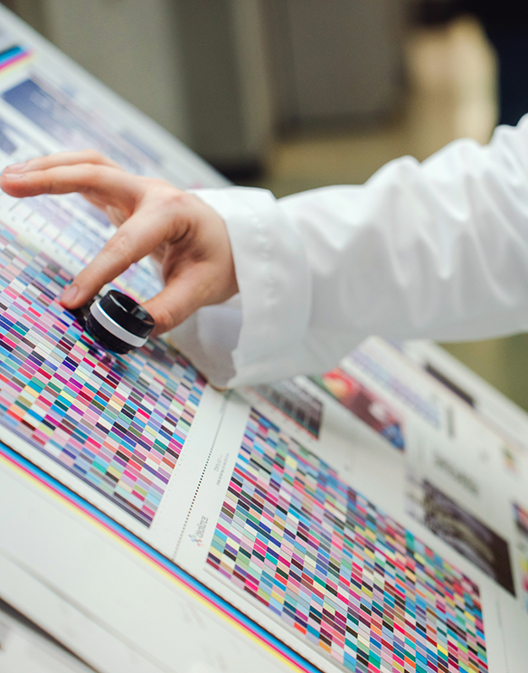 A person in a white sleeve examines color test prints on paper using a magnifying tool, checking for print quality and color accuracy.