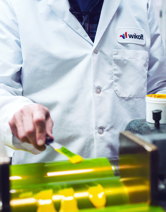 Person in a white lab coat labeled "Wikoff" applies yellow ink to a roller in an industrial printing or manufacturing setting.