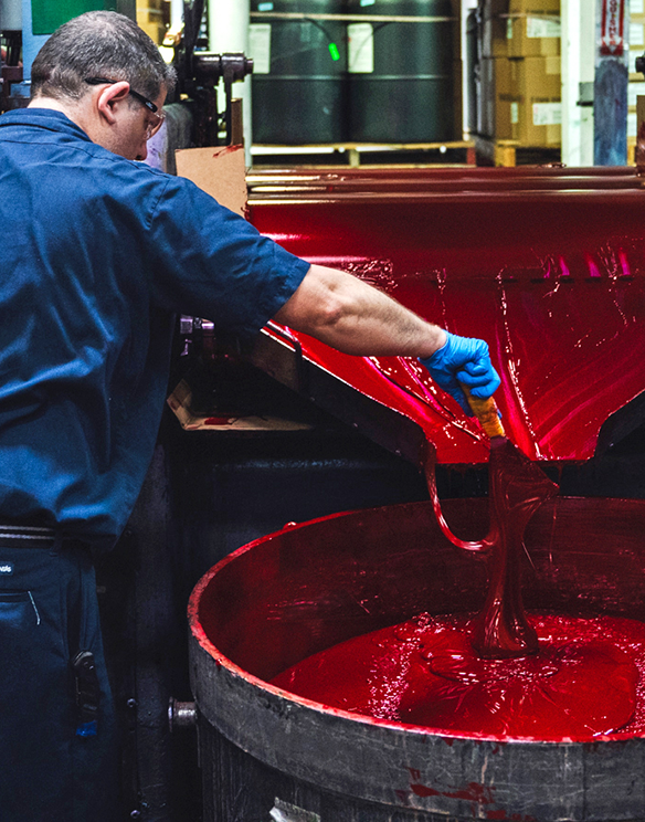 A worker in blue gloves guides thick red liquid from a machine into a large container in an industrial setting.