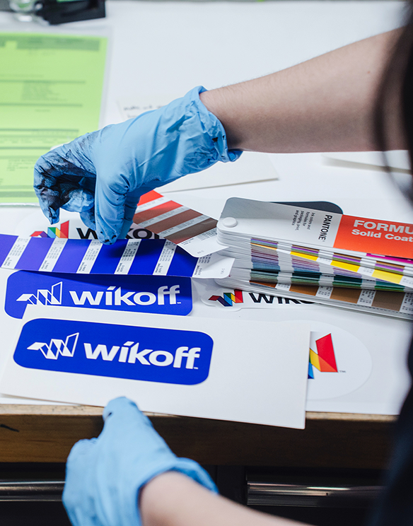 A person wearing blue gloves handles printed color samples and logo stickers labeled "wikoff" on a desk, with a Pantone color guide nearby.