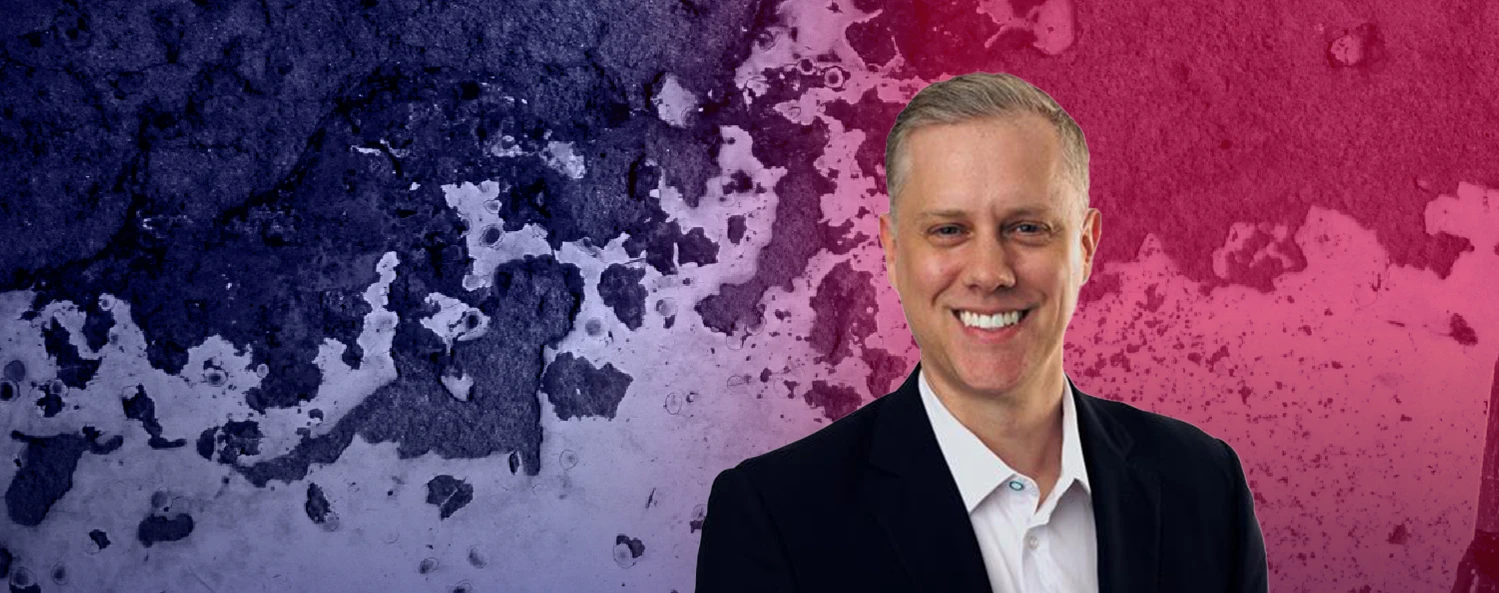 Smiling man in a suit and white shirt stands in front of a textured, multicolored background blending dark blue, purple, and pink.