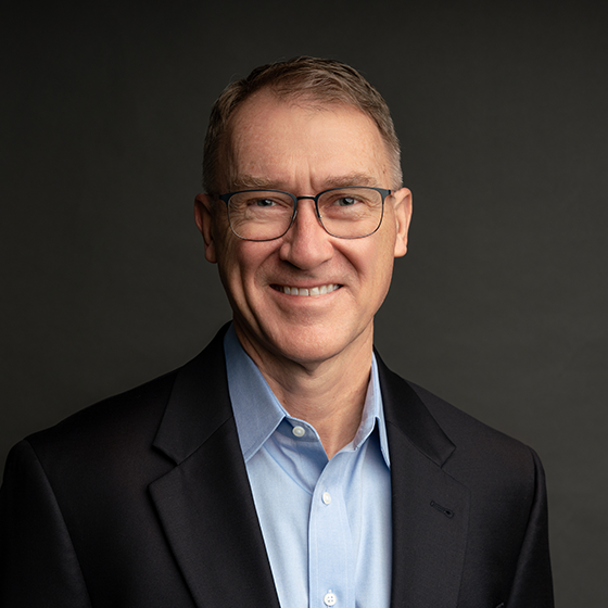 A middle-aged man wearing glasses, a light blue shirt, and a dark blazer smiles in front of a plain dark background.