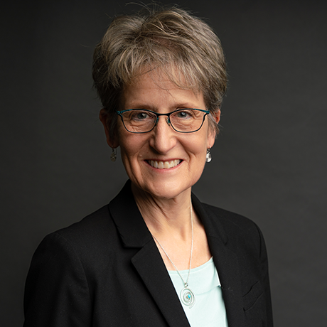 Smiling older woman with short gray hair, glasses, and a black blazer over a light top, posing against a dark background.
