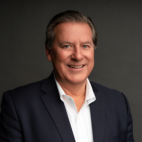 Man in a dark suit jacket and white shirt smiling against a plain dark background.