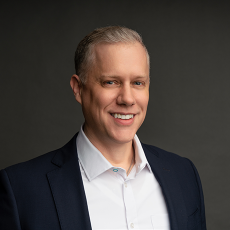 A man with short light hair wearing a dark suit jacket and white shirt, smiling, posed against a plain dark background.