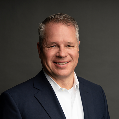 A middle-aged man in a navy suit and white shirt smiles at the camera against a plain dark background.