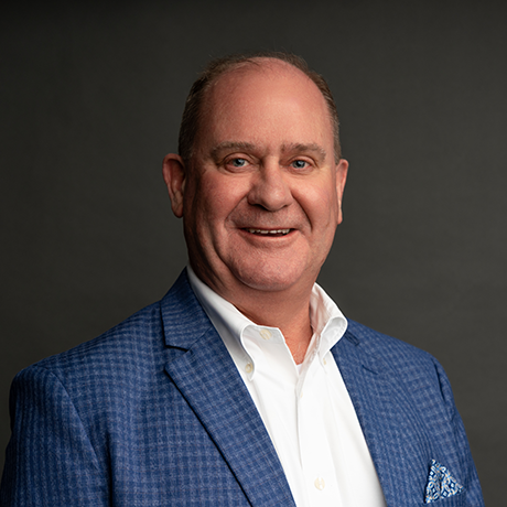 Man in a blue checkered blazer and white shirt smiles at the camera against a plain dark background.