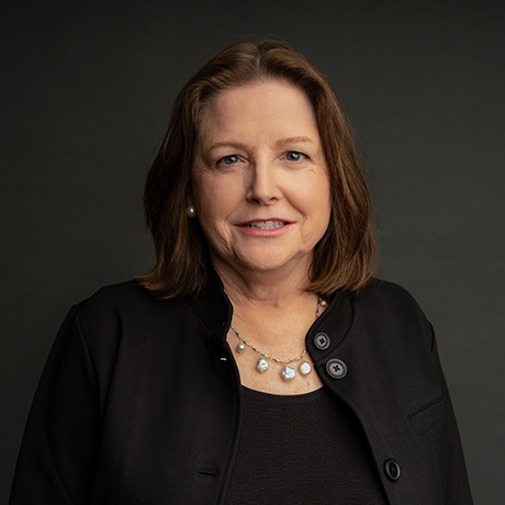 A woman with shoulder-length brown hair wearing a black jacket, a black top, and a pearl necklace poses against a dark background.