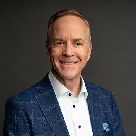 A middle-aged man wearing a blue plaid blazer and white shirt smiles at the camera against a plain dark background.