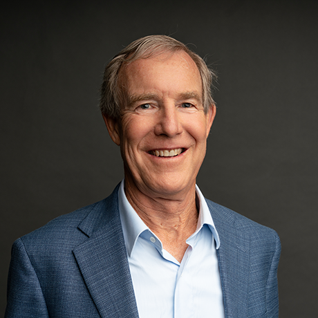 An older man with gray hair, wearing a light blue collared shirt and a blue blazer, smiles in front of a plain dark background.