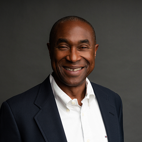A man in a dark suit jacket and white shirt smiles in front of a plain, dark background.