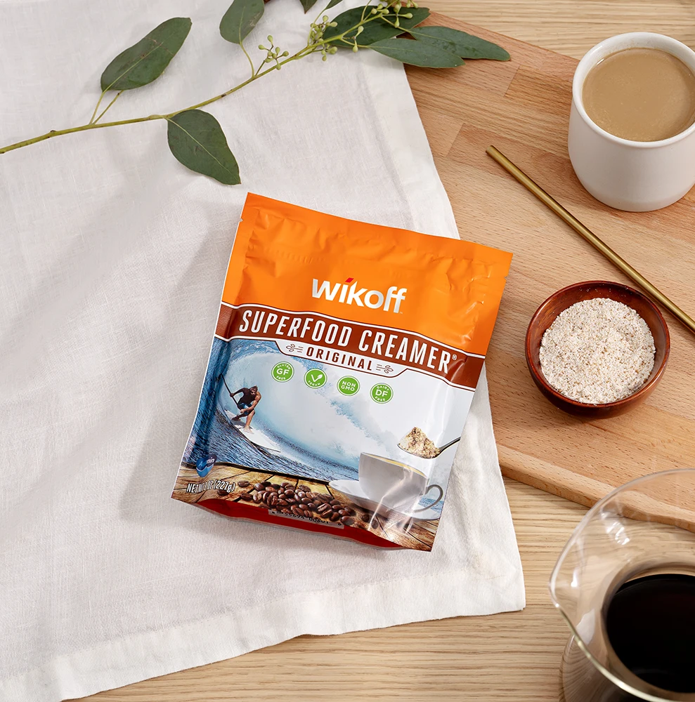 A bag of Wikoff Superfood Creamer sits on a white cloth next to a coffee cup, a glass carafe, a bowl of powder, a gold spoon, and eucalyptus leaves.