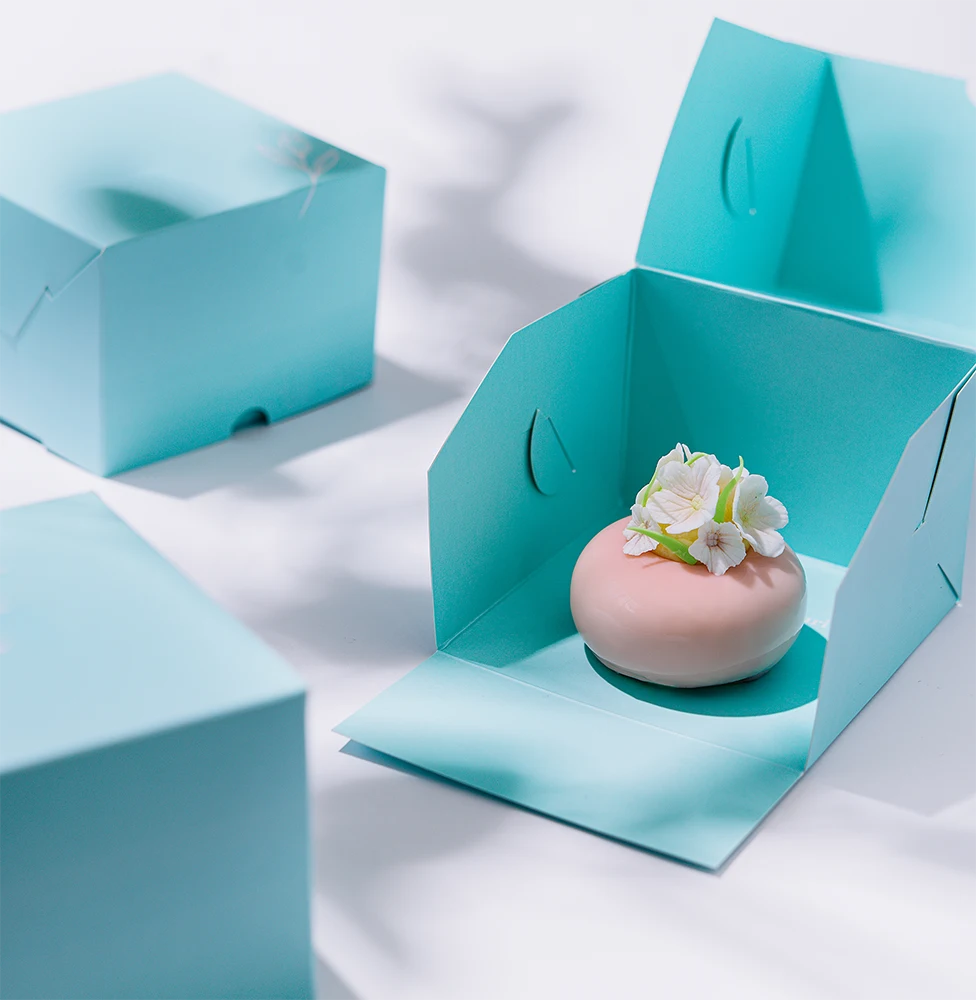 A pink glazed pastry with white flowers sits inside an open light blue box, with two closed boxes nearby on a white surface.