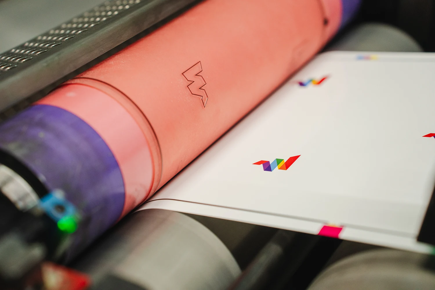 Close-up of a printing press in operation, with colored rollers transferring a multicolored geometric design onto sheets of white paper.