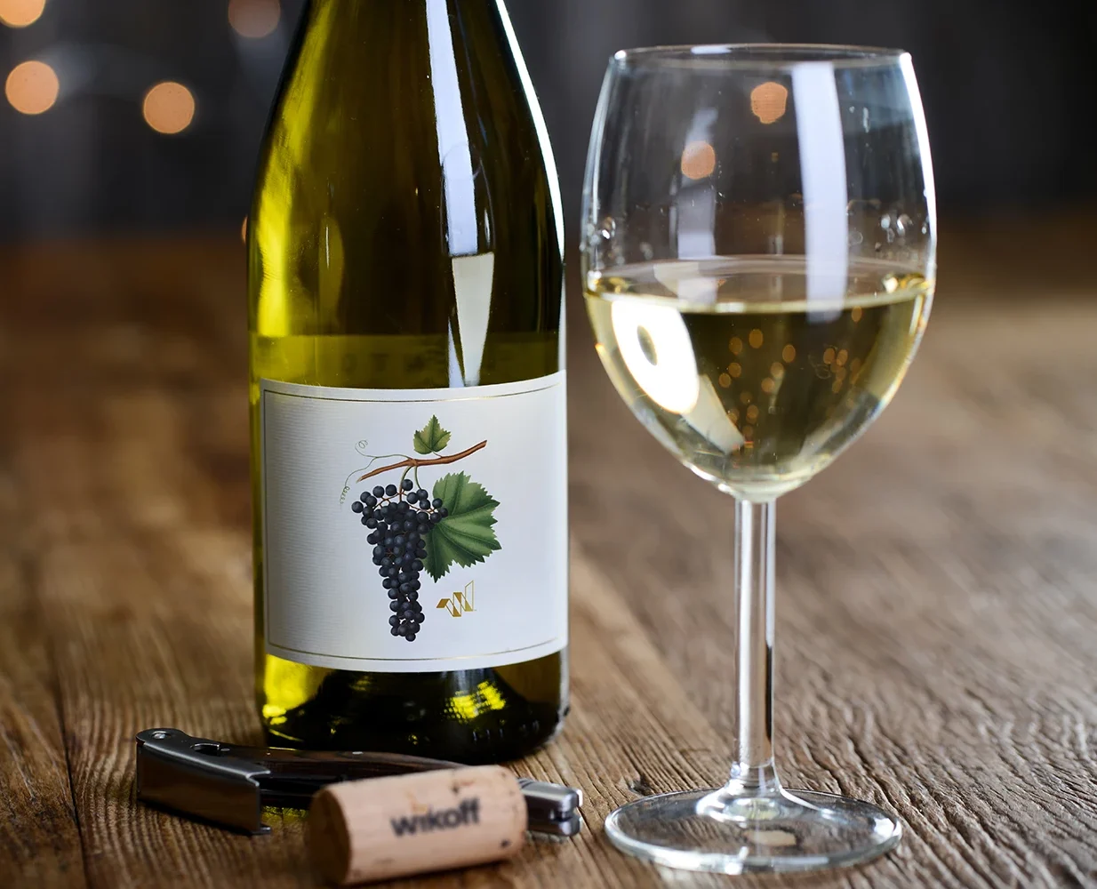 A bottle of white wine, a filled wine glass, a corkscrew, and a cork labeled "winok" on a wooden table with blurred lights in the background.