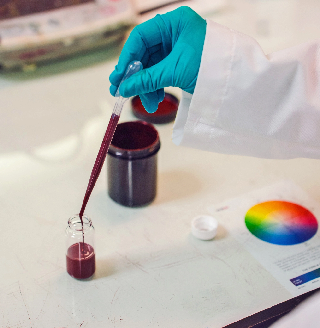 A person wearing gloves uses a pipette to transfer a dark red liquid into a small glass vial on a laboratory table with a color wheel chart nearby.
