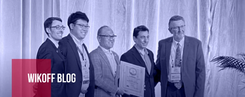 Five men in suits stand together on a stage, one holding a framed certificate or plaque, with a curtained backdrop and a “Wikoff Blog” label in the corner.