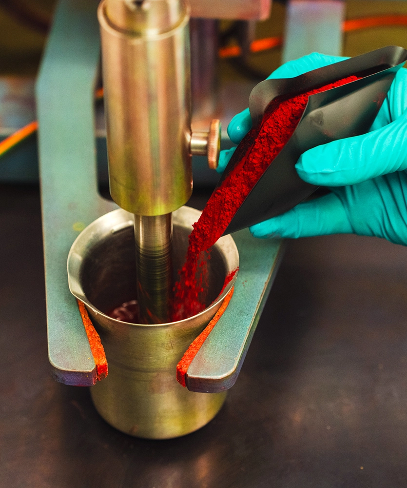 A gloved hand pours red powder from a black container into a metal beaker under laboratory equipment.