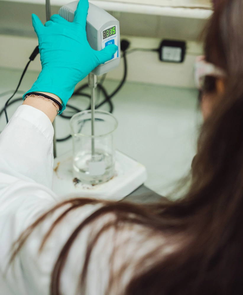 Person wearing a lab coat and teal glove operates a laboratory mixer, stirring a clear liquid in a glass beaker on a white workbench.