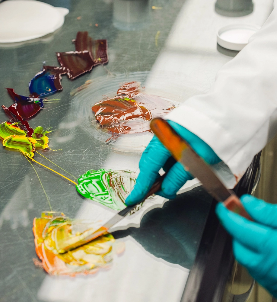 A person in blue gloves uses a spatula to mix colorful paints on a reflective surface in a laboratory or studio setting.