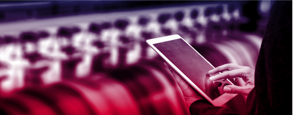 A person uses a tablet in front of industrial machinery with a blurred background, showing interaction with technology in a manufacturing or industrial setting.
