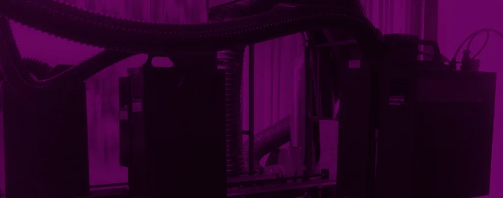 Industrial machinery with large flexible hoses and metal components, set in an indoor environment. The image has a magenta color overlay.