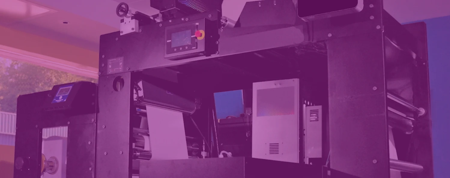 A large industrial printing machine with control panels and rollers, set in a brightly lit room with windows.